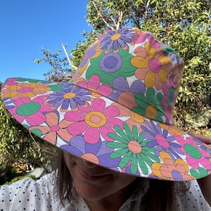 May include: A wide-brimmed sun hat with a colorful floral pattern in shades of pink, green, purple, and orange. The hat is made of fabric and has a white background.