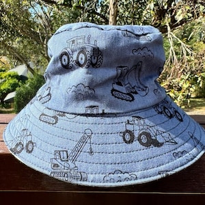 May include: A blue bucket hat with a construction vehicle print. The hat features a variety of vehicles including tractors, excavators, and cranes.