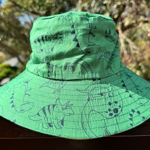 May include: Green bucket hat with a dinosaur print. The hat has a wide brim and a flat crown. The dinosaurs are black and white and include a triceratops, a stegosaurus, and a pterodactyl.