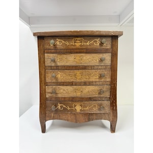 Op de afbeelding: Een kleine, antieke houten ladekast met zes laden. De kast is bruin met decoratieve inlegontwerpen en kleine, ronde knoppen. De laden hebben sierlijke bloemen- en urnenpatronen. De kast heeft een gebogen voet en staat tegen een witte achtergrond.