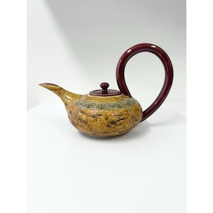 May include: A decorative teapot with a brown and yellow mottled finish. The teapot features a dark red handle and lid, and a spout. The body of the teapot has a geometric design around the middle.
