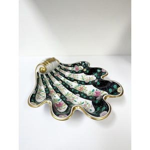 May include: A decorative, shell-shaped dish with a gold rim and accents. The dish features a black, white, and green striped pattern with floral designs. The dish has a gold-colored, scroll-like handle.