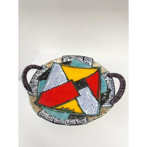 Op de afbeelding: Een kleurrijk, abstract keramisch serveerblad met twee donkerbruine handvatten. De lade heeft een geometrisch ontwerp met rode, gele, blauwe en grijze vormen. De rand heeft een decoratief patroon.