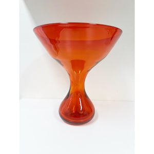 Può includere: Un vaso di vetro arancione vibrante con una parte superiore svasata e una base arrotondata. Il vaso ha una finitura liscia e lucida e un'esclusiva forma a clessidra. Questo pezzo decorativo è ideale per esporre fiori o come oggetto d'arte autonomo.