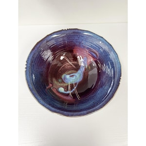 Puede incluir: Un cuenco de cerámica hecho a mano con un acabado brillante. El cuenco presenta un borde azul oscuro que se desvanece en un centro burdeos intenso. El interior tiene un diseño único con acentos azules y verdes.