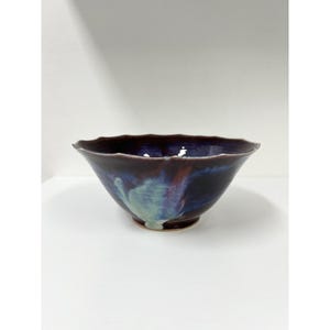 Puede incluir: Un cuenco de cerámica con exterior morado oscuro y borde ondulado. El cuenco presenta una mezcla de tonos azules, verdes y rojos, creando una apariencia artística única. El cuenco es adecuado para servir alimentos o como pieza decorativa.