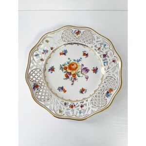 May include: A white porcelain plate with a scalloped edge and a gold rim. The plate features a floral design in the centre with orange, purple, and blue flowers. The outer edge has a lattice design.