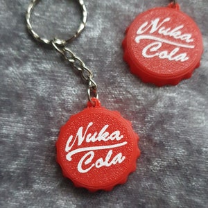 May include: Two red bottle cap keychains with the white text "Nuka Cola" on them. The keychains are on a grey textured surface.