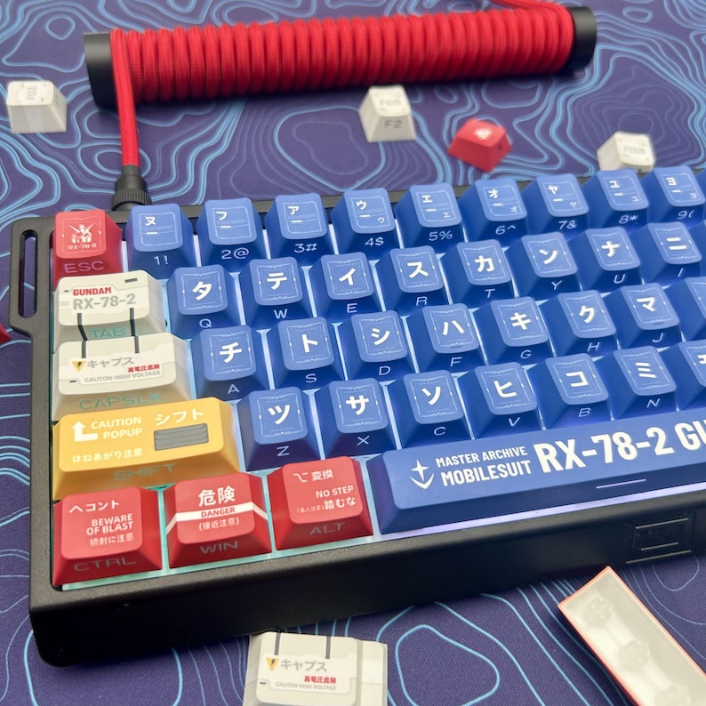Puede incluir: Un teclado mec&aacute;nico azul y blanco con caracteres japoneses y etiquetas en ingl&eacute;s. El teclado tiene teclas de acento rojas y amarillas, y un cable en espiral rojo. El teclado est&aacute; etiquetado como "RX-78-2" y "MOBILESUIT".