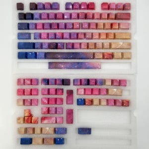 Puede incluir: Un conjunto de teclas de teclado personalizadas en una variedad de colores, incluyendo tonos de rosa, morado, azul y marr&oacute;n. Las teclas tienen un dise&ntilde;o tem&aacute;tico de galaxia, con una apariencia estrellada. Las teclas est&aacute;n dispuestas en un inserto de espuma blanca.