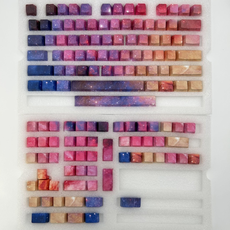 Puede incluir: Un conjunto de teclas de teclado personalizadas en una variedad de colores, incluyendo tonos de rosa, morado, azul y marr&oacute;n. Las teclas tienen un dise&ntilde;o tem&aacute;tico de galaxia, con una apariencia estrellada. Las teclas est&aacute;n dispuestas en un inserto de espuma blanca.