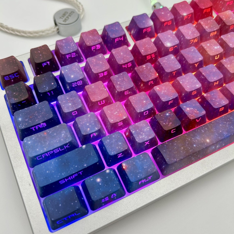 Puede incluir: Primer plano de un teclado mec&aacute;nico con teclas personalizadas. Las teclas presentan un dise&ntilde;o de galaxia en tonos azules, morados y rojos. El teclado tiene un marco plateado y est&aacute; iluminado con un brillo azul y morado. Las teclas est&aacute;n etiquetadas con letras blancas.