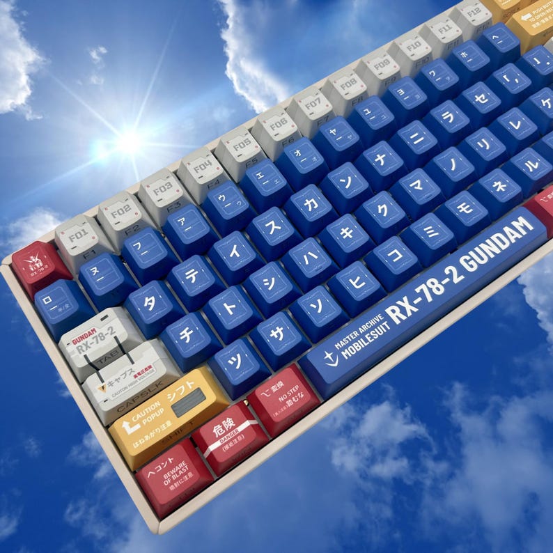 Puede incluir: Un teclado mec&aacute;nico con teclas azules y blancas, con caracteres japoneses y texto en ingl&eacute;s. El teclado tiene un tema Gundam, con el texto "RX-78-2 GUNDAM" en la barra espaciadora. Otras teclas tienen texto japon&eacute;s y etiquetas en ingl&eacute;s.
