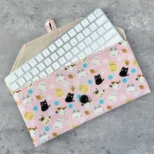 May include: A pink fabric pouch with a repeating pattern of cats and balls of yarn. A white wireless keyboard is partially visible inside the pouch.