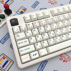May include: Off-white mechanical keyboard with Japanese and English characters. The keyboard has a retro gaming theme, with colorful keycaps and a matching joystick. The keyboard has a 1 Player key and a Control key.