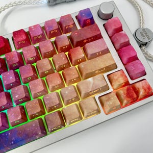 May include: A close-up of a silver mechanical keyboard with custom keycaps. The keycaps feature a galaxy-like design in shades of pink, red, orange, and purple. The keyboard has a silver frame and a volume knob. The keycaps have white lettering.