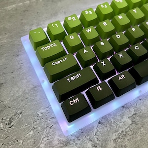 May include: A green and black mechanical keyboard with white keycaps and RGB lighting. The keyboard has a gradient color scheme, with the top row of keys being green and the bottom row being black. The keyboard is illuminated with a white light.