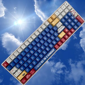Puede incluir: Un teclado mec&aacute;nico personalizado con una combinaci&oacute;n de colores azul, blanco, rojo y amarillo. Las teclas presentan caracteres japoneses y letras inglesas. El teclado tiene el texto "RX-78-2 GUNDAM" en el lateral.