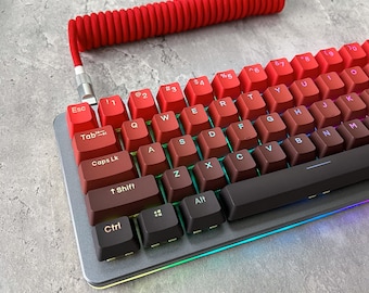 May include: A red and black mechanical keyboard with a red coiled cable. The keyboard has a rainbow backlight and features keys with white lettering. The keys are labelled with letters, numbers, and symbols.