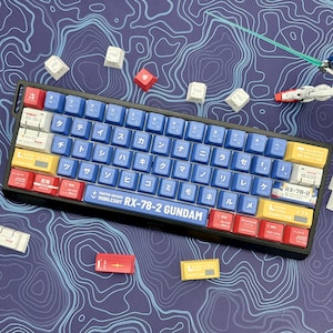 Puede incluir: Un teclado mec&aacute;nico negro con teclas azules, rojas y amarillas. El teclado tiene caracteres japoneses y el texto "RX-78-2 GUNDAM". Teclas adicionales est&aacute;n dispersas sobre un mapa topogr&aacute;fico azul.