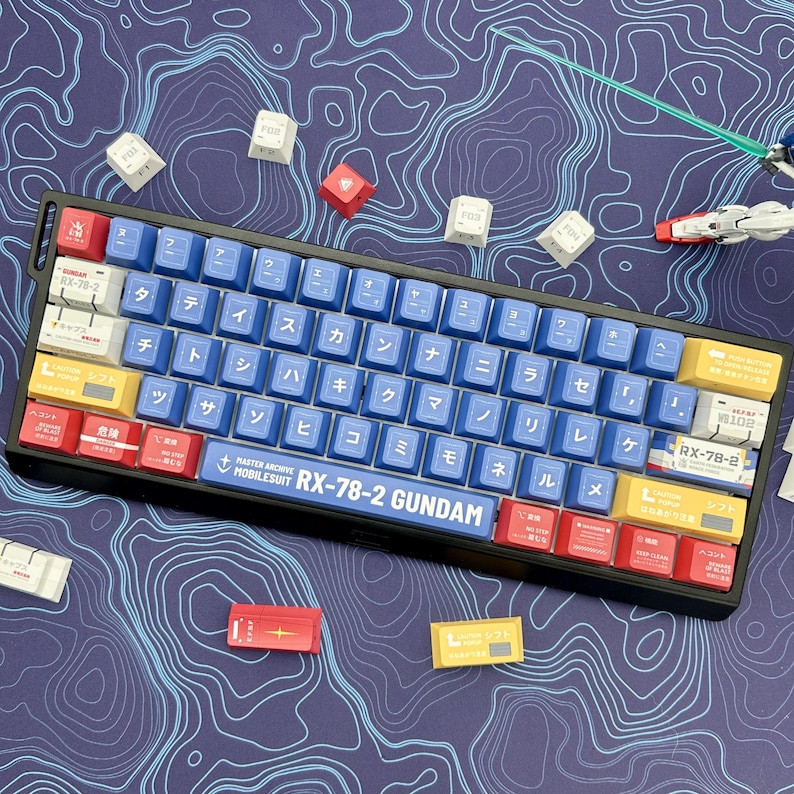 Puede incluir: Un teclado mec&aacute;nico negro con teclas azules, rojas y amarillas. El teclado tiene caracteres japoneses y el texto "RX-78-2 GUNDAM". Teclas adicionales est&aacute;n dispersas sobre un mapa topogr&aacute;fico azul.