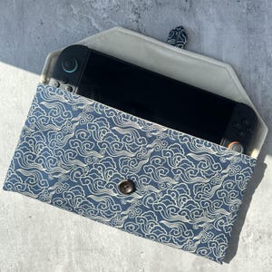 May include: A blue and white patterned case with a button closure, holding a black handheld gaming device. The case features a cloud-like design and a neutral-colored interior. The button is brown.