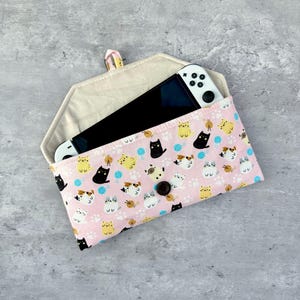 May include: A pink fabric case with a cat pattern, holding a white and black handheld gaming device. The case features a button closure and a beige interior. The cat pattern includes black, yellow, and calico cats with yarn balls and paw prints.
