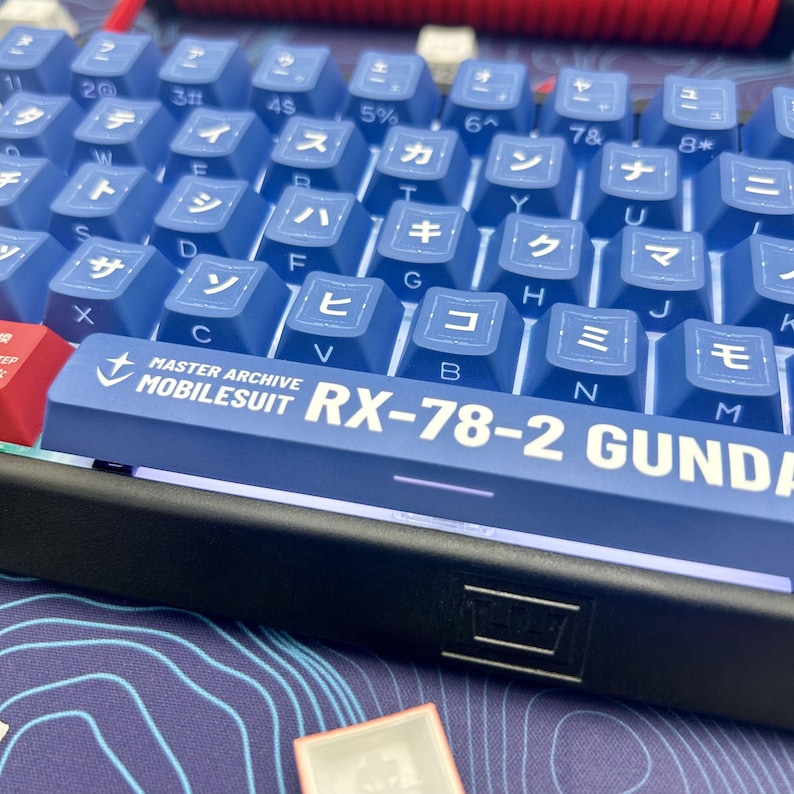 Puede incluir: Primer plano de un teclado mec&aacute;nico con teclas azules y caracteres japoneses. La barra espaciadora presenta el texto "MASTER ARCHIVE MOBILESUIT RX-78-2 GUNDAM" en blanco. El teclado tiene un marco negro y un cable rojo.