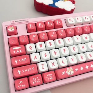 May include: A pink mechanical keyboard with red and white keycaps. The keycaps feature symbols, letters, and a Shiba Inu dog illustration. The keyboard has a 2.4GHz RF transceiver.