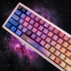 Puede incluir: Un teclado mec&aacute;nico de oro rosa con teclas con un dise&ntilde;o de galaxia colorido. Las teclas cambian de azul a rosa a naranja, con un patr&oacute;n de estrellas. El fondo es una escena espacial oscura con una nebulosa.