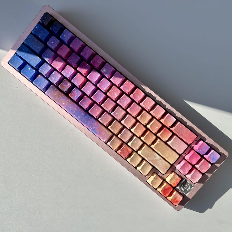 Puede incluir: Un teclado mec&aacute;nico compacto de color oro rosa con teclas con un dise&ntilde;o de galaxia colorido. Las teclas cambian de azul a rosa, naranja y amarillo. El teclado tiene una distribuci&oacute;n QWERTY est&aacute;ndar.