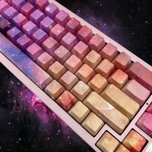 Puede incluir: Primer plano de un teclado mec&aacute;nico con teclas con un dise&ntilde;o de galaxia en tonos de rosa, morado y naranja. El teclado tiene un marco rosa claro y un fondo tem&aacute;tico espacial.