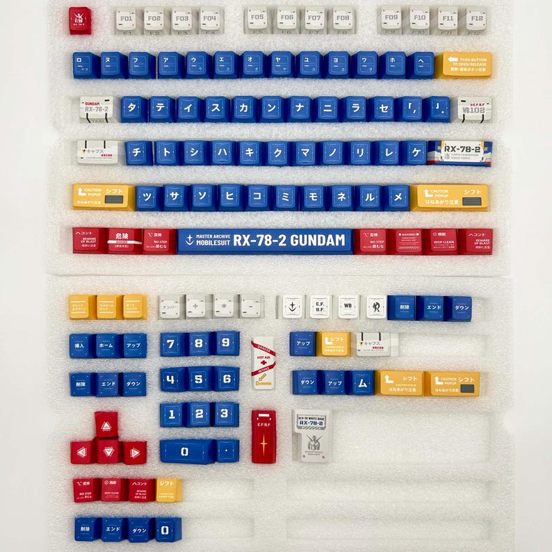 Puede incluir: Un conjunto de teclas de teclado mec&aacute;nico de colores, incluyendo azul, amarillo, rojo y blanco. Algunas teclas presentan caracteres japoneses y el texto "RX-78-2 GUNDAM". Las teclas est&aacute;n dispuestas en una bandeja.