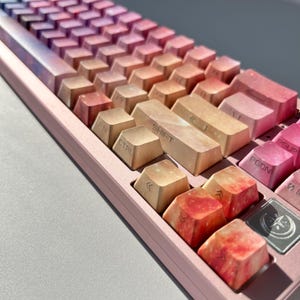 Puede incluir: Primer plano de un teclado mec&aacute;nico con teclas con un dise&ntilde;o degradado de tonos rosa, naranja y morado. El teclado tiene un marco rosa y teclas de funci&oacute;n con texto blanco. Las teclas tienen un dise&ntilde;o gal&aacute;ctico.