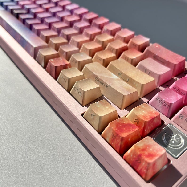 Puede incluir: Primer plano de un teclado mec&aacute;nico con teclas con un dise&ntilde;o degradado de tonos rosa, naranja y morado. El teclado tiene un marco rosa y teclas de funci&oacute;n con texto blanco. Las teclas tienen un dise&ntilde;o gal&aacute;ctico.