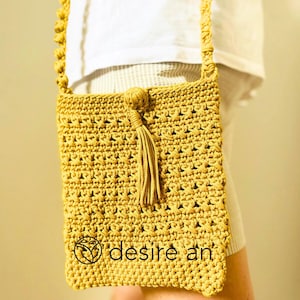 Puede incluir: Un bolso bandolera de crochet amarillo mostaza con un borla. El bolso tiene un diseño geométrico sencillo y el nombre de la marca "desire an" es visible en la parte inferior.