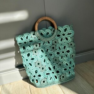 May include: A teal crocheted tote bag with a wooden handle. The bag is made of a delicate, openwork pattern.