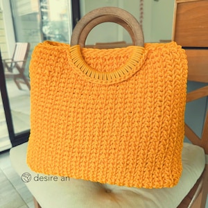 May include: A bright yellow, hand-crocheted tote bag with a wooden handle. The bag has a rectangular shape and a textured stitch pattern. The handle is made of two curved wooden pieces. The bag is sitting on a chair with a cream-colored cushion.