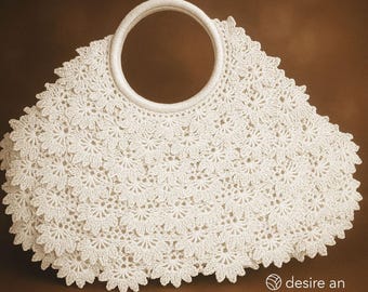 May include: A white, crocheted handbag with a circular handle. The bag features a floral pattern and a scalloped edge. The handle is also white and appears to be made of a woven material. The bag is set against a brown background.