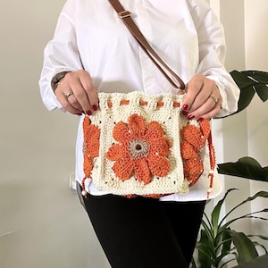 May include: A crocheted bag with a floral design in shades of orange and white. The bag has a brown leather strap and is worn over the shoulder.