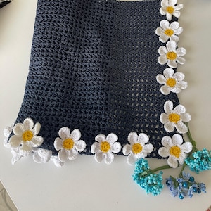 Könnte beinhalten: Eine schwarze gehäkelte Decke mit einem Rand aus weißen gehäkelten Gänseblümchen. Die Decke ist mit blauen und grünen Blumenakzenten verziert.