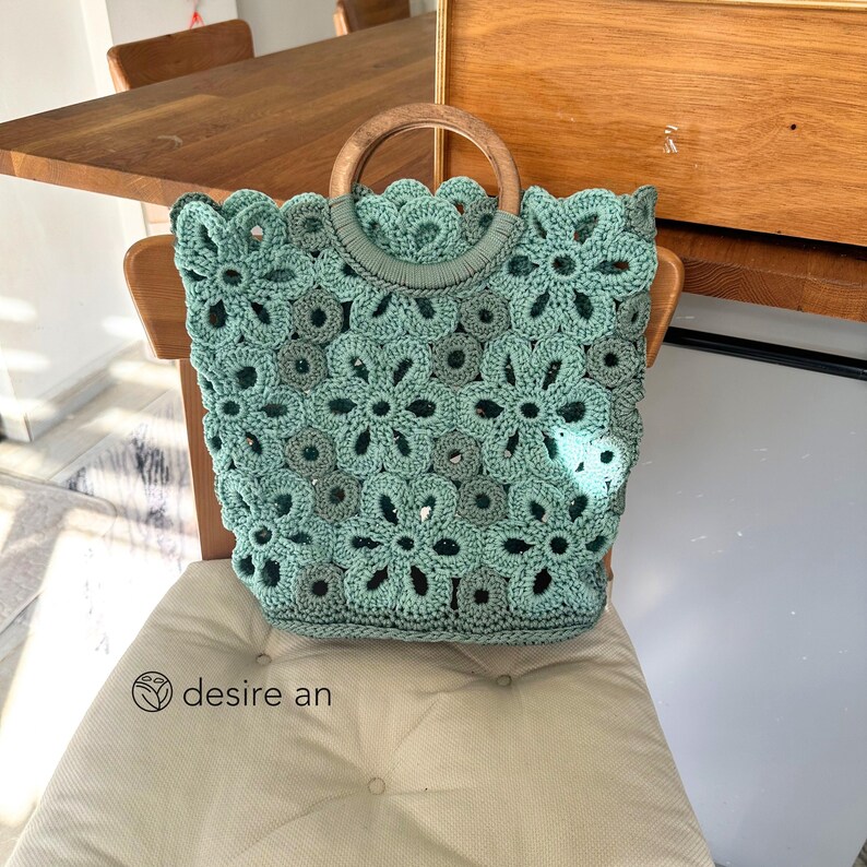 May include: A light green crocheted tote bag with a wooden handle. The bag is made of a delicate, openwork pattern and has a floral design.