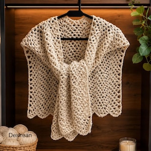 May include: A cream-coloured crochet shawl with a scalloped edge, suspended from a black hanger. The openwork pattern is evident, and the shawl is arranged to display its form. The word "Desirean" is visible in the lower left corner.