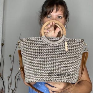 May include: A crocheted tote bag with a brown and beige color pattern. The bag has a wooden handle and a brown leather strap. The bag is held by a person.