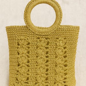 Op de afbeelding: Een mosterdgele gehaakte tote bag met een ronde handgreep. De tas heeft een bloemenpatroon op de voorkant en is gemaakt van een getextureerd garen. De tas is klein van formaat, geschikt voor het dragen van essentials.