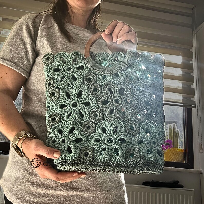 May include: A light green crocheted tote bag with a wooden handle. The bag has a floral pattern and is made with a delicate crochet stitch.