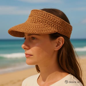 Könnte beinhalten: Gehäkelter Visor in warmem Braun. Der Visor hat eine breite Krempe und eine strukturierte, gewebte Optik. Der Hintergrund zeigt einen Strand mit blauem Wasser und klarem Himmel.