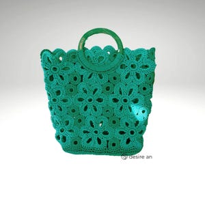 May include: A teal crocheted tote bag with a wooden handle. The bag is made of a delicate, open-weave pattern with a floral design.