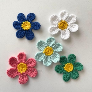 May include: Five crocheted flowers in shades of blue, white, light blue, pink, and green. Each flower features a yellow centre. Ideal for craft projects, these handmade flowers add a touch of colour.