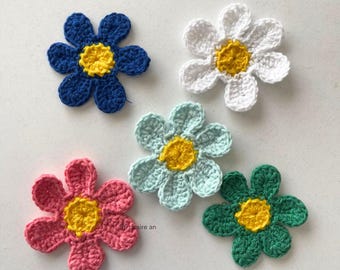 May include: Five handmade crochet flowers in various colors: blue, white, light blue, pink, and green. Each flower has a yellow center. These decorative flowers are perfect for crafting projects.